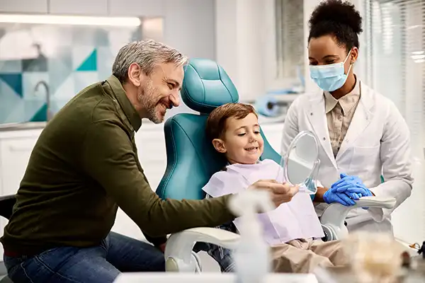 White father holding a mirror for his happy, young son sitting in a dental chair while his female dentist happily watches